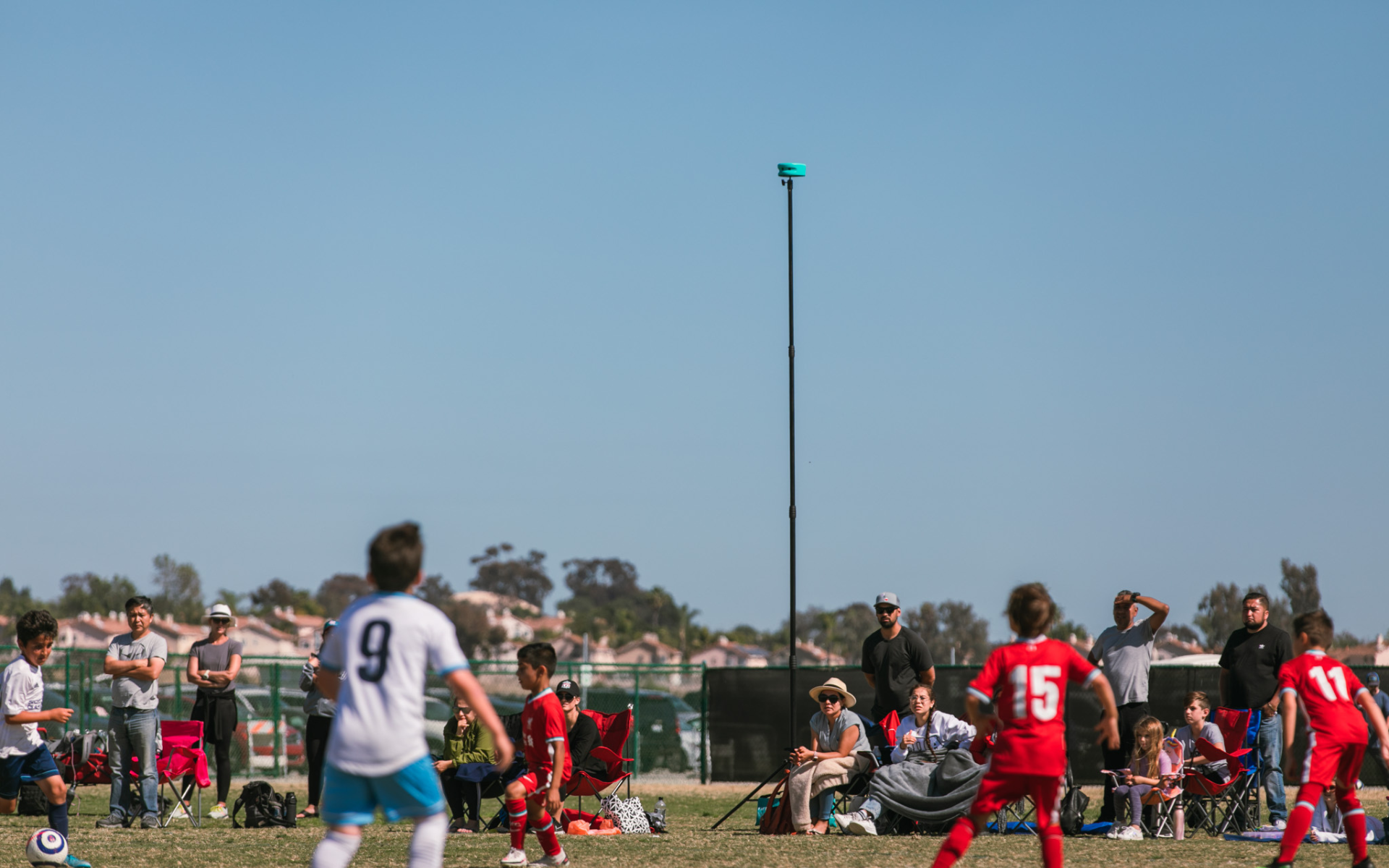 Trace camera on a tall pole above a youth soccer game with players and seated spectators