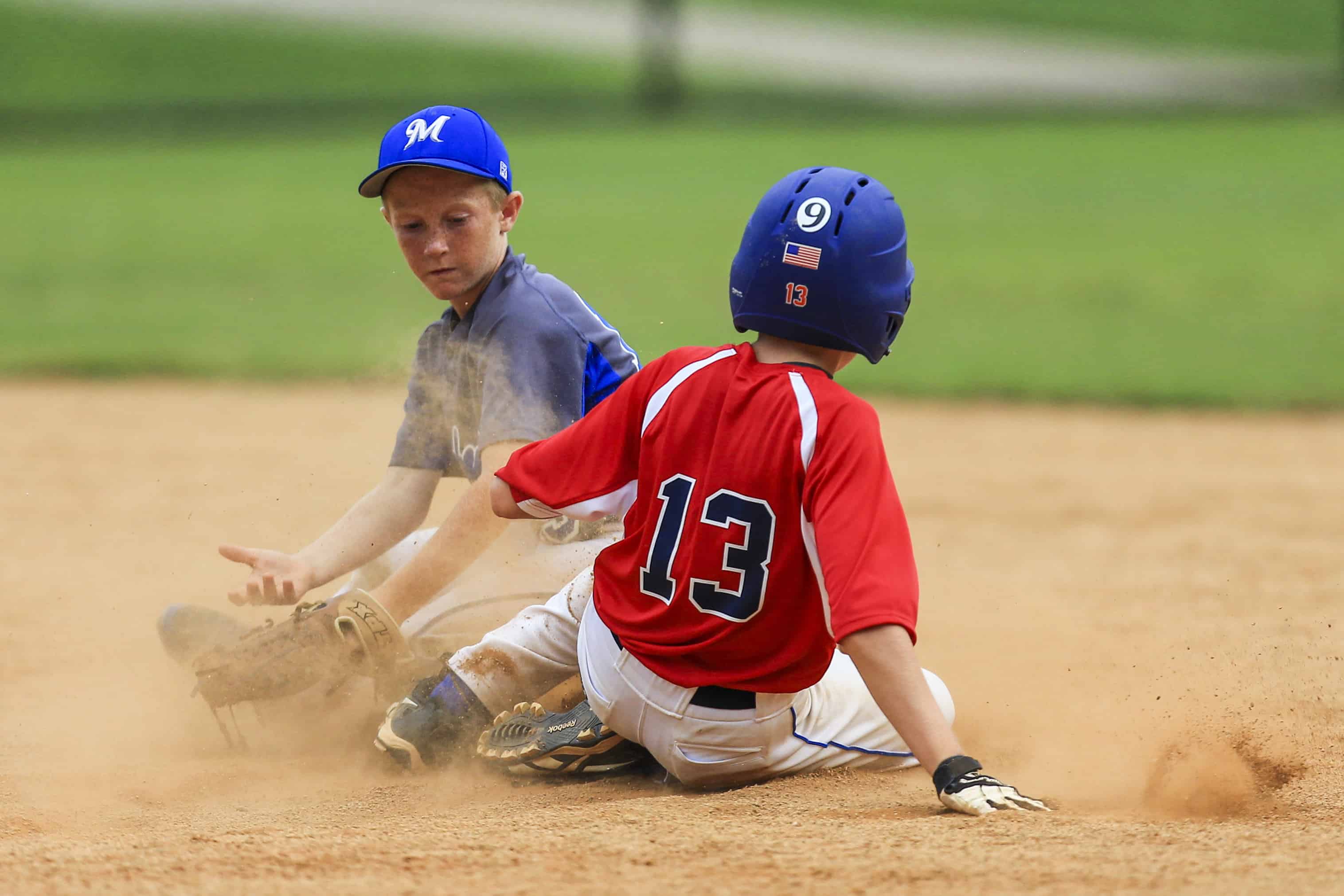 Youth baseball runner in red sliding into base in a cloud of dust as a fielder applies the tag