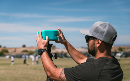 Man Mounting Trace Camera Man in a cap and sunglasses mounting a teal Trace camera phone on a tripod at a soccer field