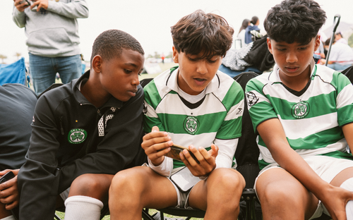 Players Watching Phone Together Three youth soccer players in green-and-white kits watching video on a phone together