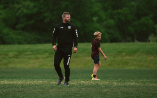 Coach in all black standing on a green field watching a young player in maroon walk by