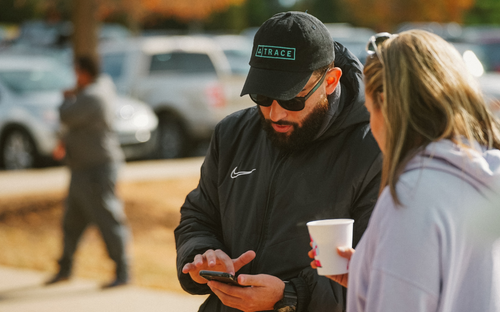 Parent Checking Trace App Man in a black Trace cap and sunglasses looking at his phone beside a woman, holding a coffee cup outdoors