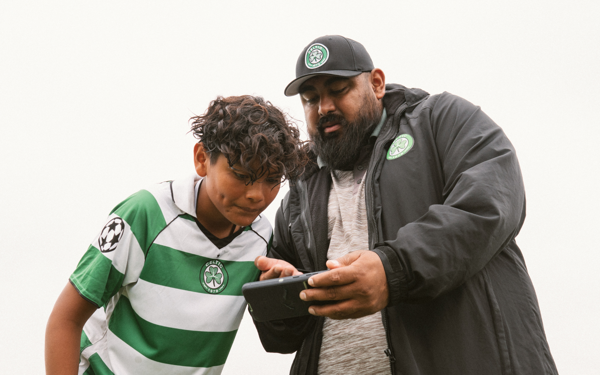 Coach Reviewing Video With Player Bearded coach in a Celtic cap and jacket showing phone footage to a young player in a green-and-white hooped jersey