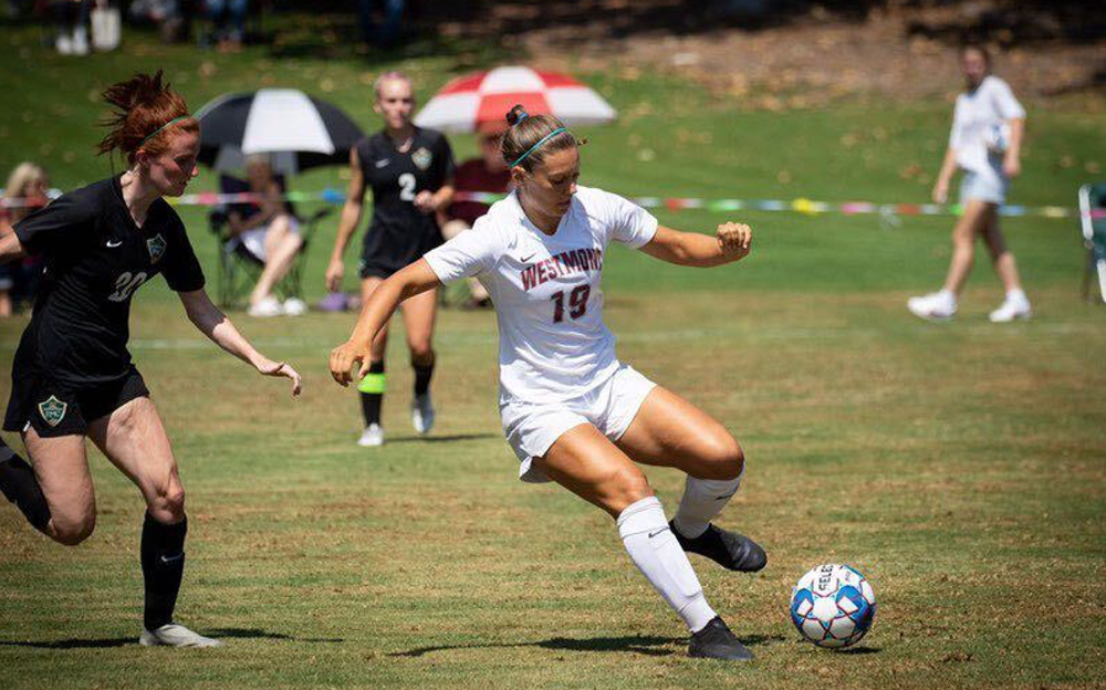Female Westmont player number 19 in white shielding the ball from a defender in black during a sunny match