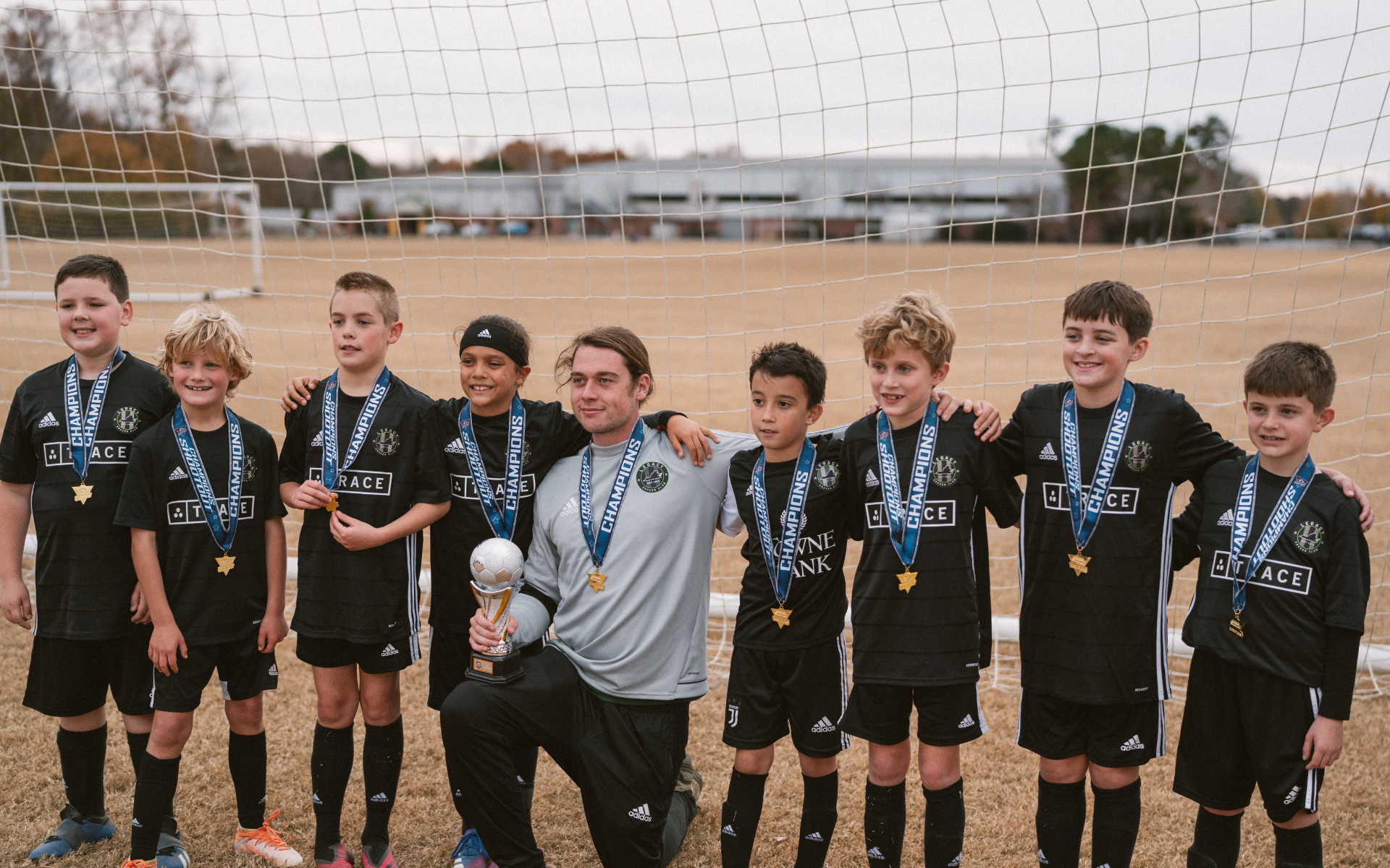 Youth Team With Championship Trophy Youth boys soccer team in black Trace kits wearing medals and holding a trophy in front of the goal with coach kneeling