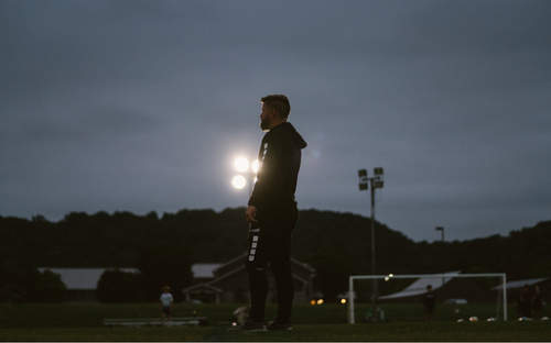 Silhouette of a coach standing alone on a soccer field at dusk with stadium lights glowing behind
