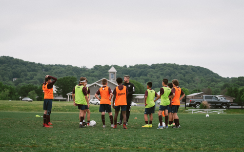 Coach addressing a circle of youth players in orange training bibs on a field with green hills behind
