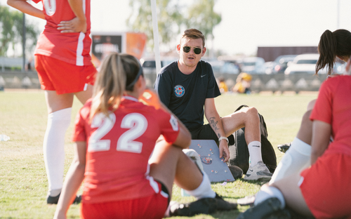 Coach in sunglasses seated on the field with a tactics board talking to female players in red gathered around him