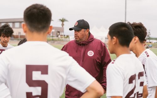 Coach in a maroon jacket and cap speaking to players in white and maroon jerseys on the field