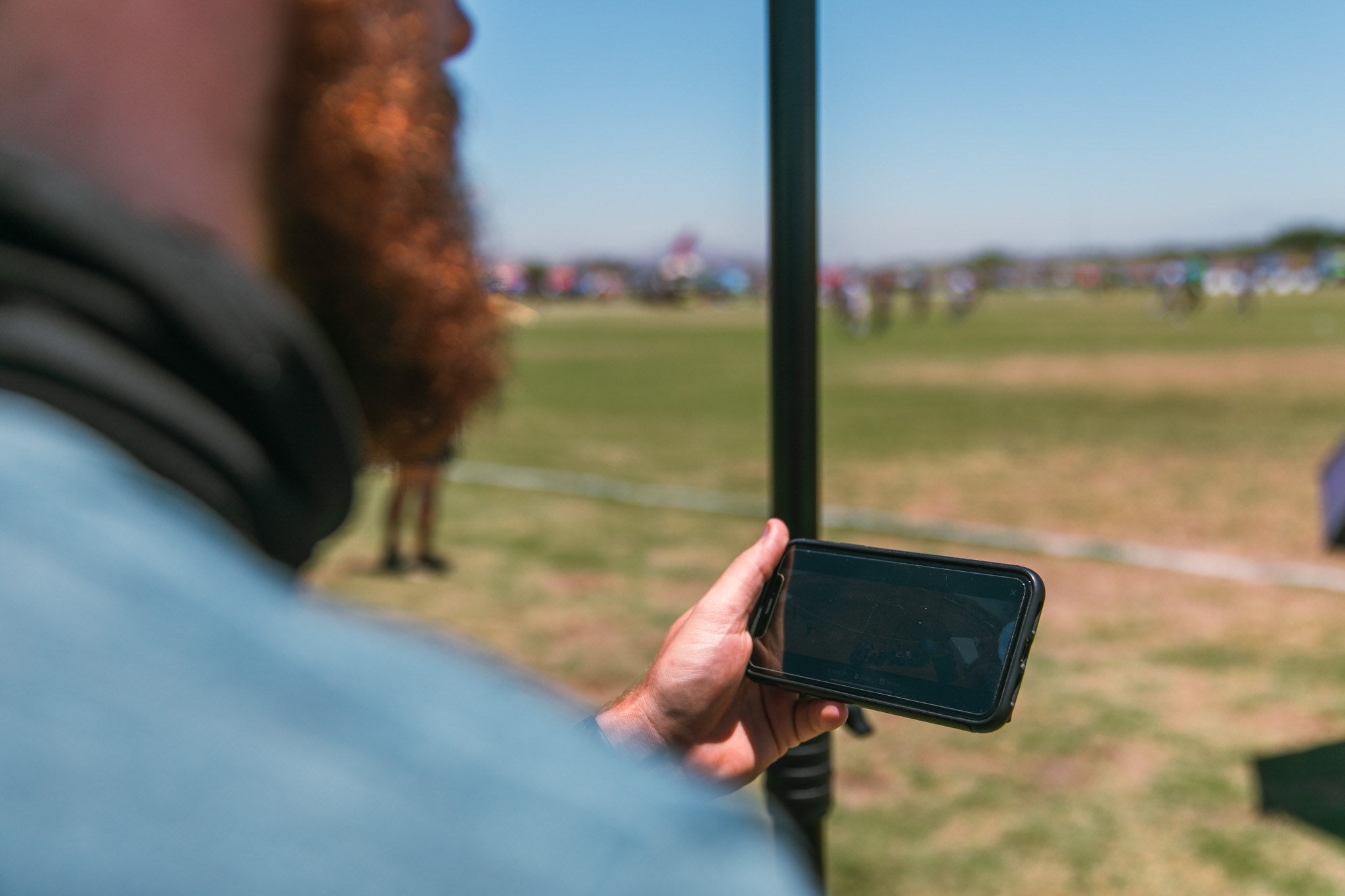 Filming A Game By Phone Over-the-shoulder view of a bearded man holding a smartphone while watching a distant soccer field