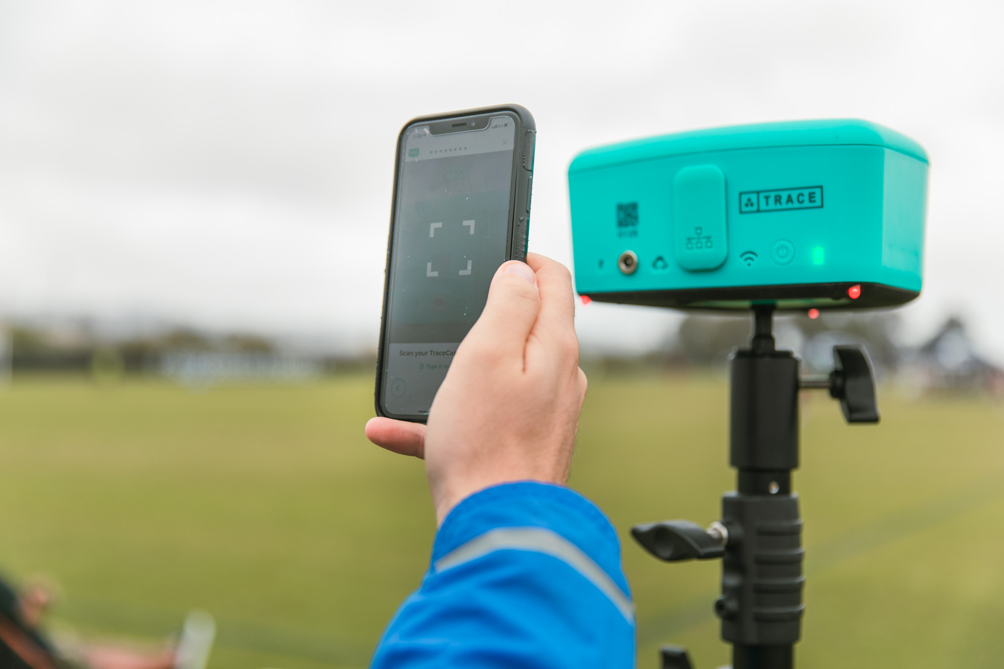 Hand holding a phone scanning a QR code on a teal Trace camera mounted on a tripod at a field