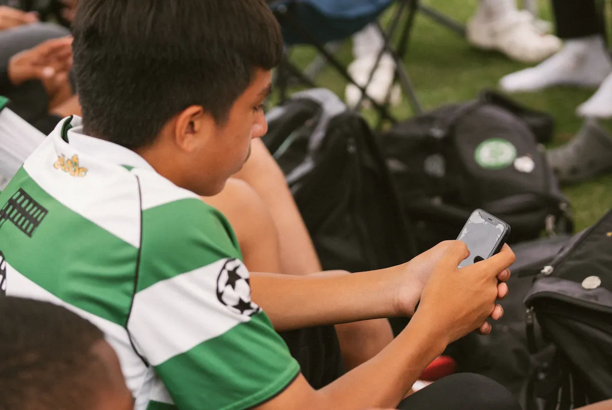 Boy in a green-and-white hooped jersey looking at his smartphone while sitting on the grass beside kit bags