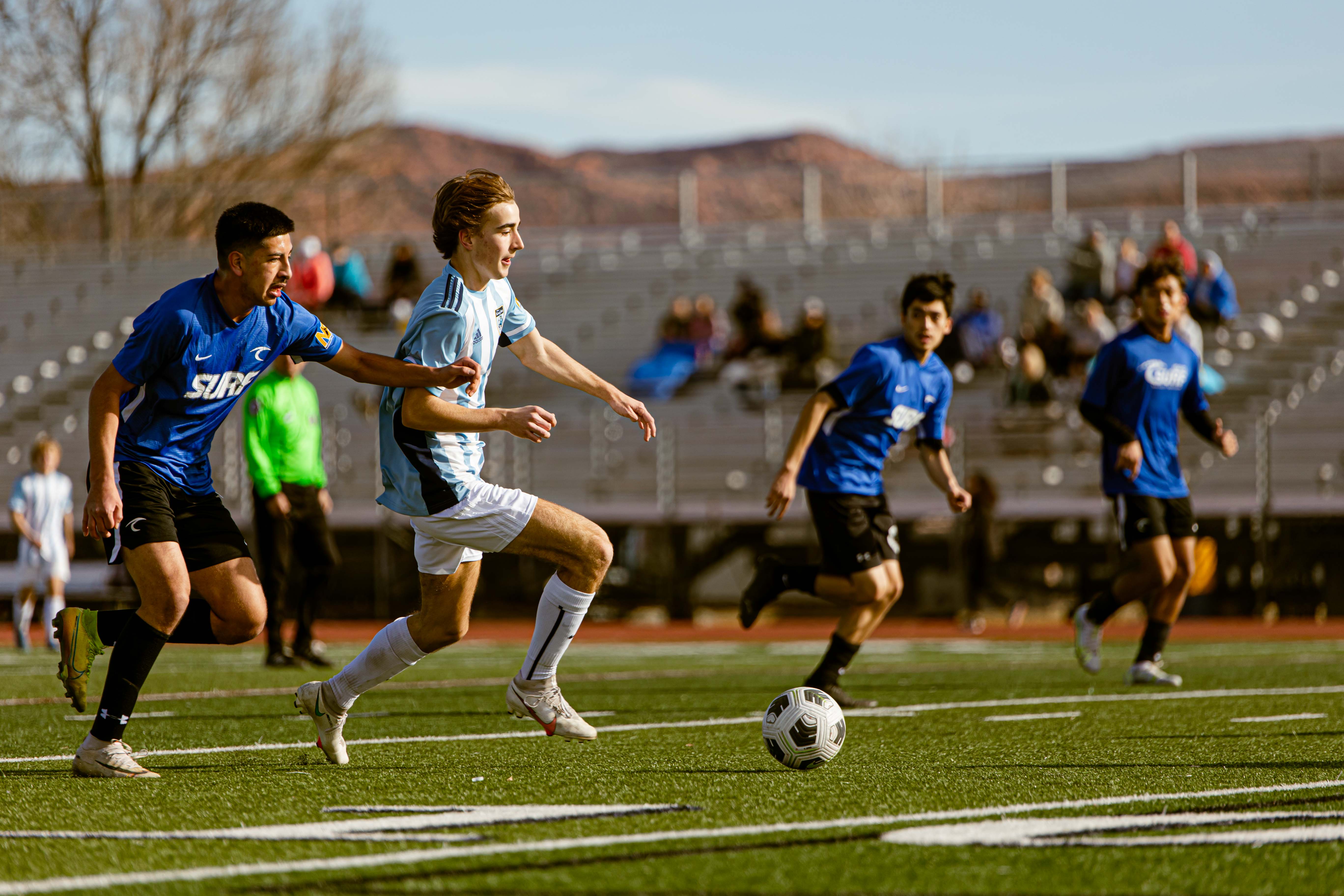 Boy in a light blue kit driving the ball forward past blue Surf defenders in a stadium match