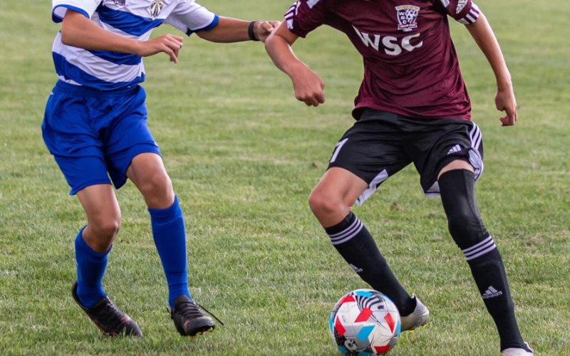 Two youth players competing for the ball, one in a blue kit and one in a maroon WSC jersey