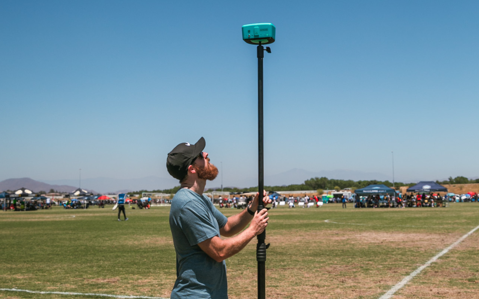 Man in a cap raising a Trace camera on a tall pole at a sunny soccer complex