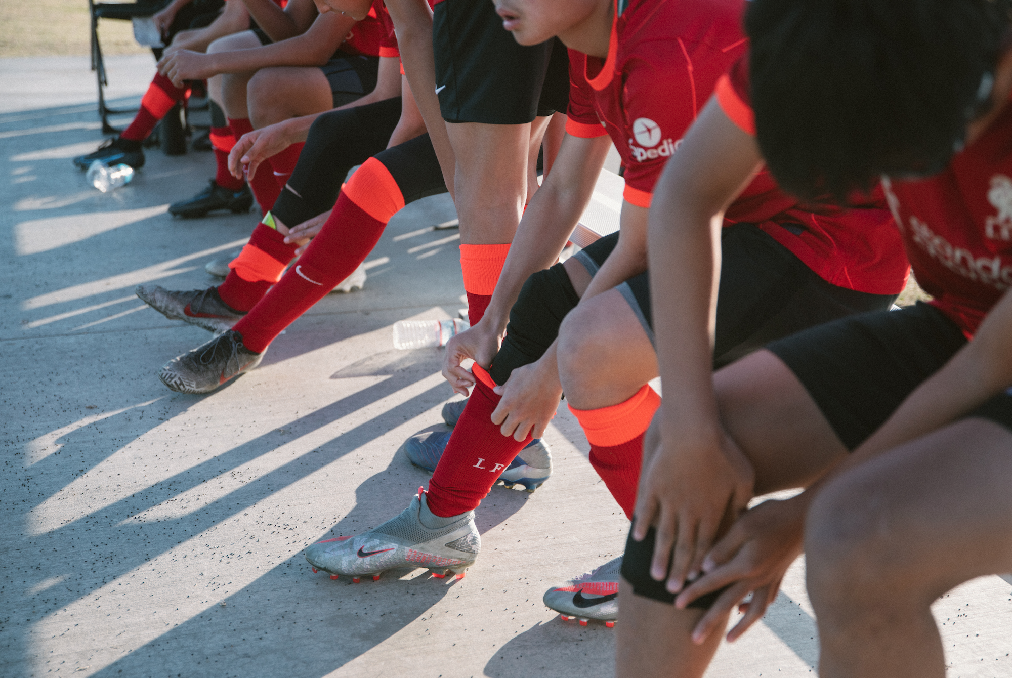 Row of youth players in red Liverpool kits sitting on a curb, close on legs and cleats