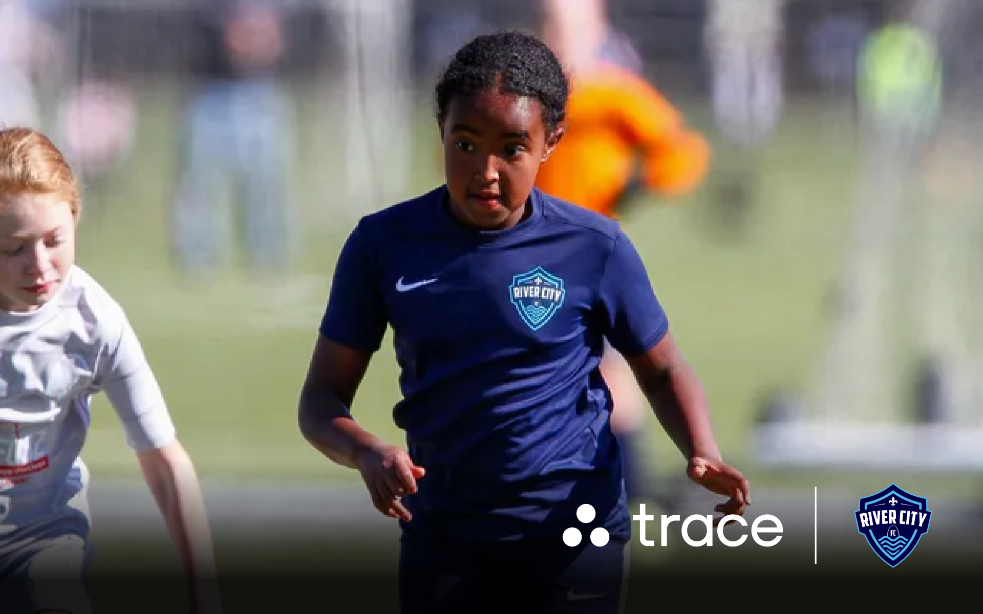 River City Player Running Girl in a navy River City kit running with the ball past an opponent, with Trace logos