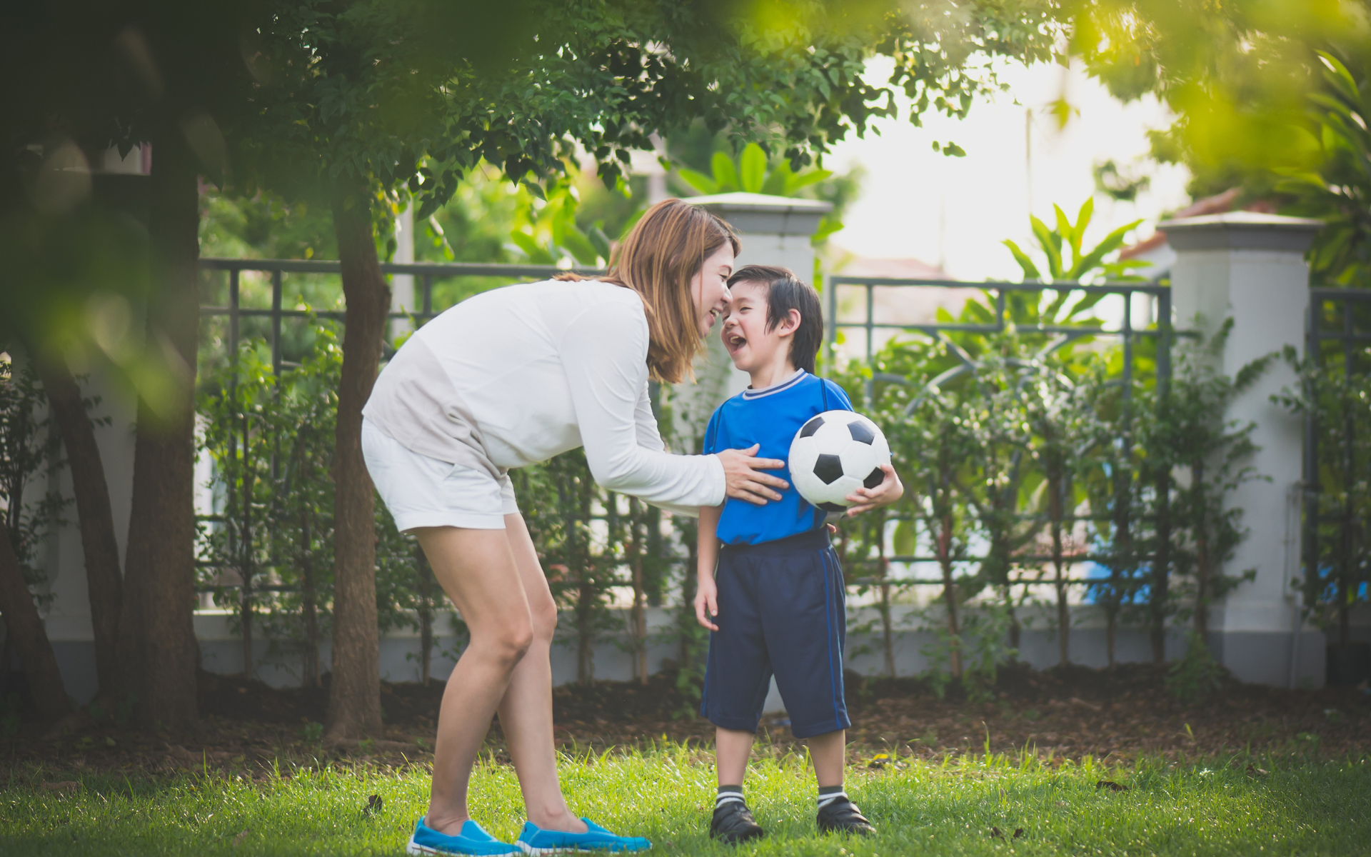 Mother bending down smiling with a young boy holding a soccer ball in a garden