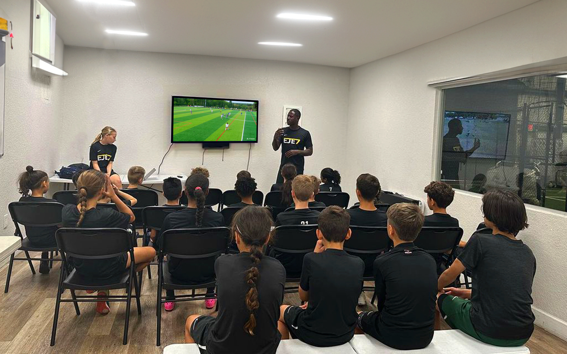Player Workshop With Kids Coach in EJE7 gear speaking to seated youth players watching soccer on a TV indoors