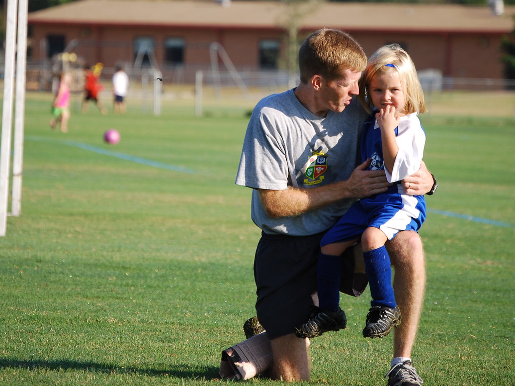 Dad kneeling on a field holding his young daughter in a blue soccer uniform