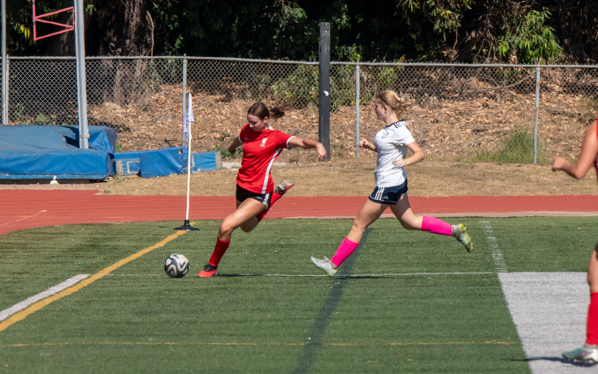 Girls Soccer Match Action Girl in red jersey striking the ball near the corner flag as a white-jersey defender chases.
