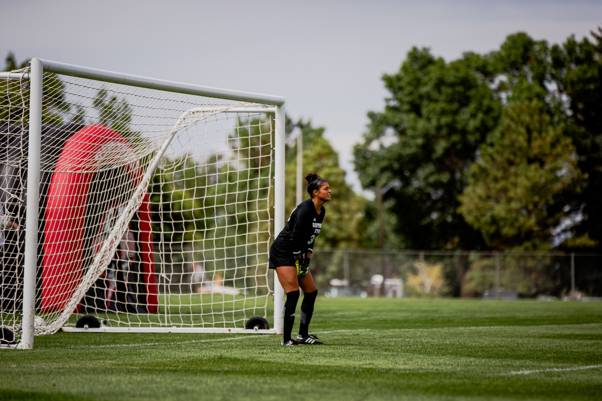 Goalkeeper Guarding The Net Female goalkeeper in black standing ready in front of the goal net with trees behind.