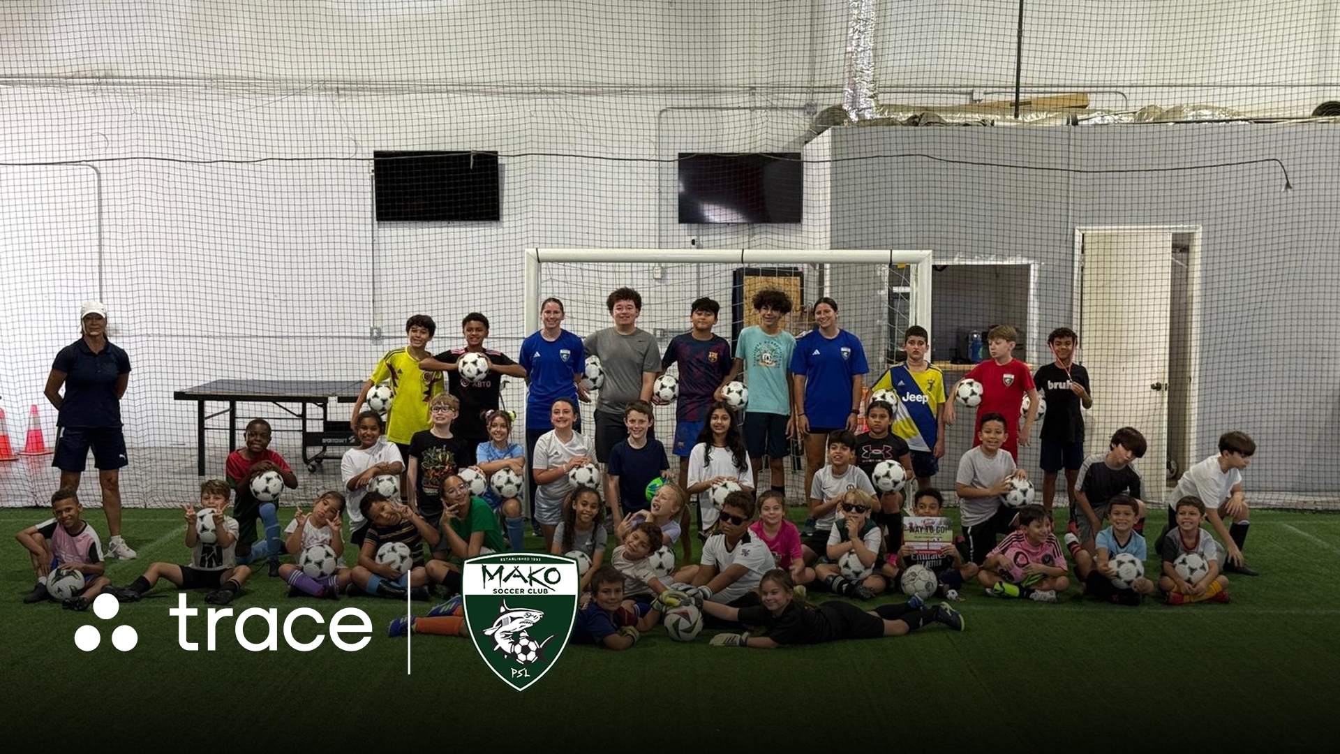 Large group of youth soccer players posing with balls at an indoor facility, with Trace and Mako logos.